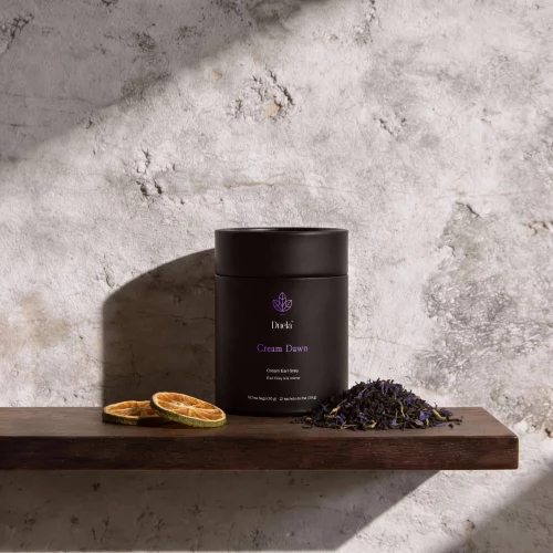 Duela Cream Dawn black tea in a matte black tube, photographed on a dark wooden shelf against a textured stone wall with dried orange slices and loose black tea in soft natural light.